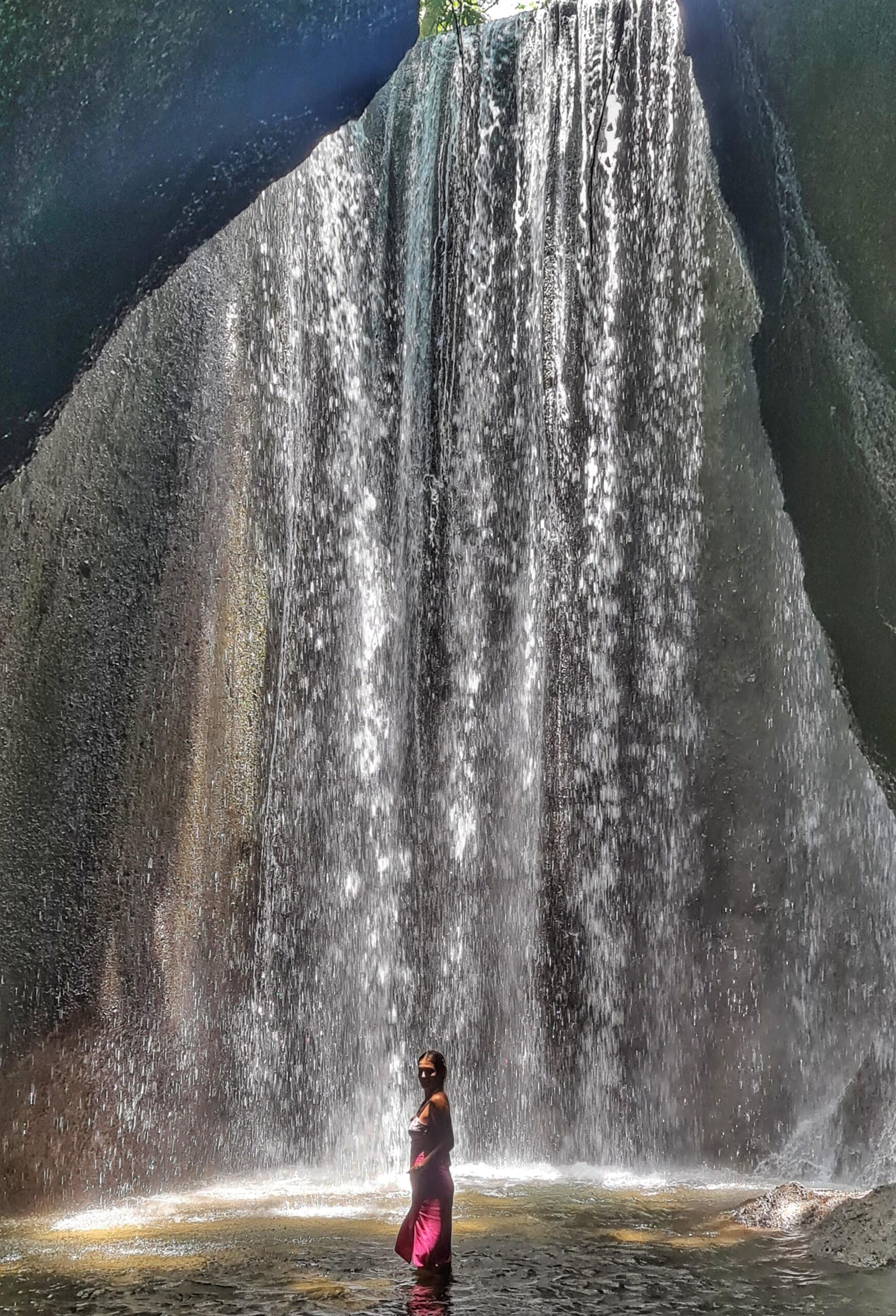 Traveler standing beside a Bali waterfall, looking up at the cascading water and towering cliffs.