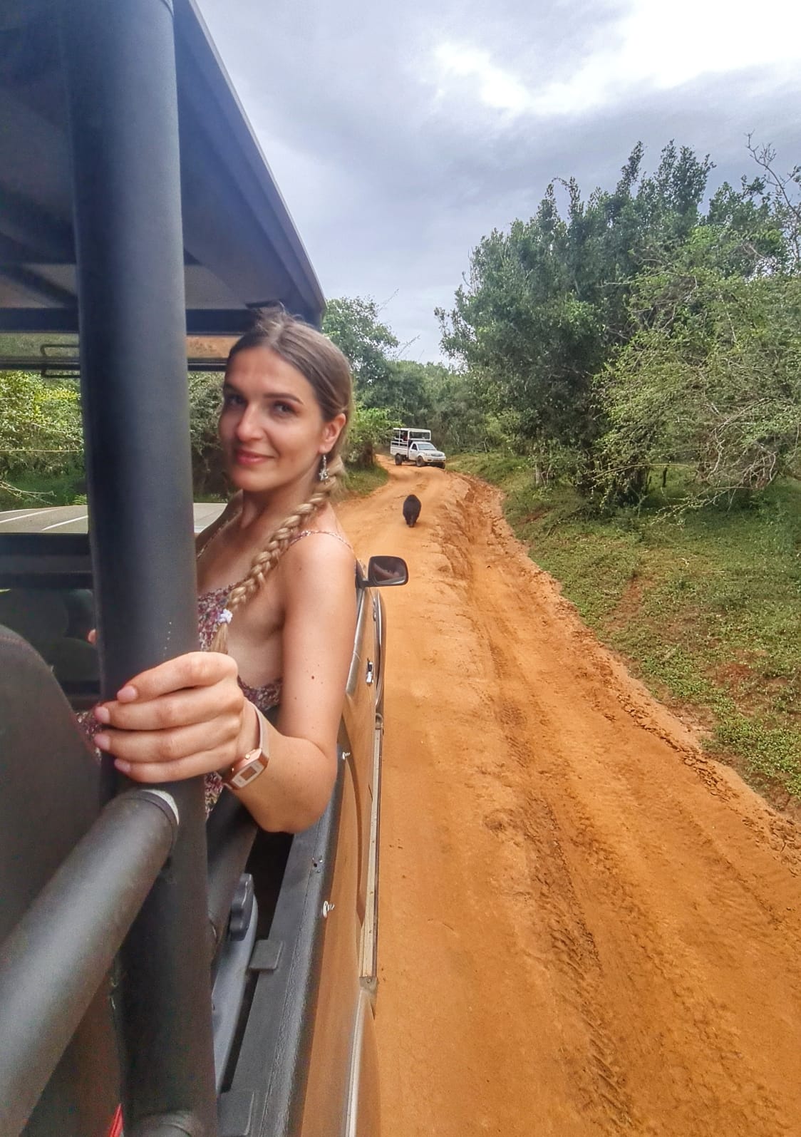 Woman enjoying a safari ride on a dirt road with wildlife and lush greenery around.
