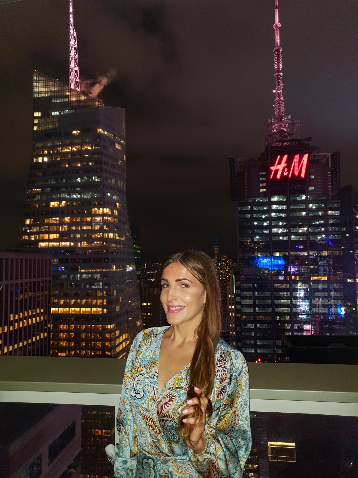 Person standing at a rooftop bar in Midtown Manhattan at night with city skyline behind.
