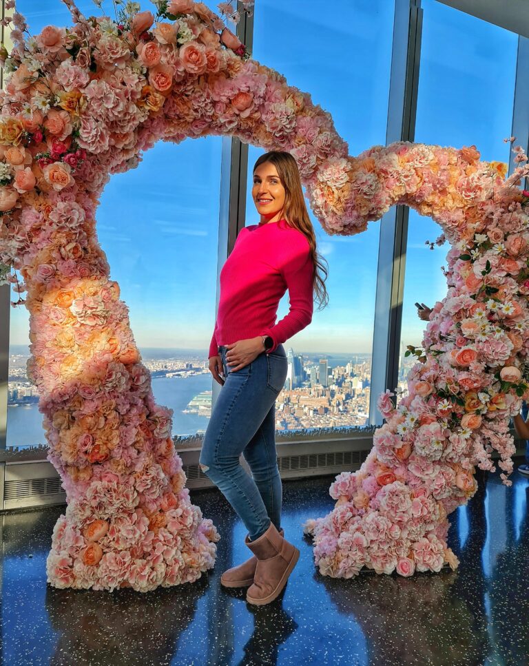 Standing at One World Observatory with heart-shaped frame and New York City skyline in the background